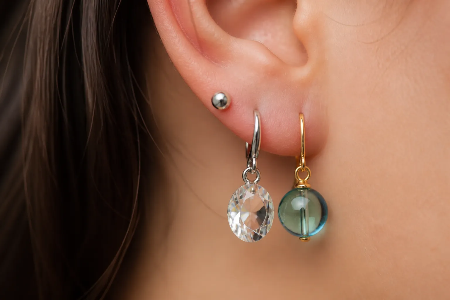 sterling silver earrings - Close-up editorial photo comparing a sterling silver stud earring, a rhodium-plated brass crystal drop earring, and a gradient glass bead earring side by side on a human earlobe model, illustrating visual difference in finish, scale, and light interaction