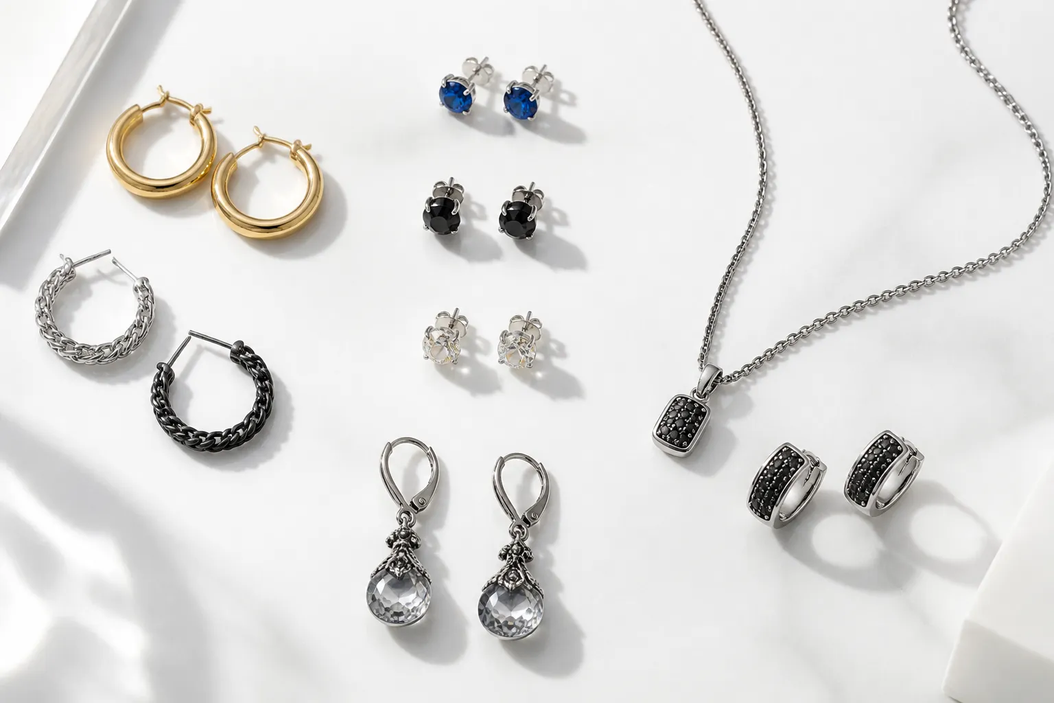 pendientes hombre - Fotografía de producto plana (flat lay) mostrando una selección variada de pendientes hombre: aros, studs con piedra, pendientes de gota con cristal vintage y set pendiente más collar, sobre fondo blanco limpio con etiquetas de estilo
