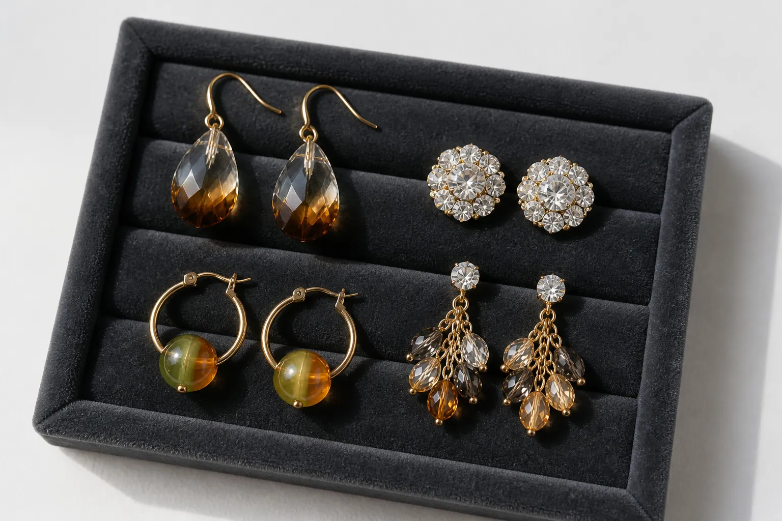 pendientes mujer - Fotografía editorial sobre fondo blanco limpio mostrando cuatro pares de pendientes mujer de diferentes estilos —colgantes de cristal degradado, botones de pedrería, aros con cuentas de vidrio bicolor y conjuntos con cadena— dispuestos en filas ordenadas sobre terciopelo gris oscuro, luz natural lateral que resalta los reflejos del cristal