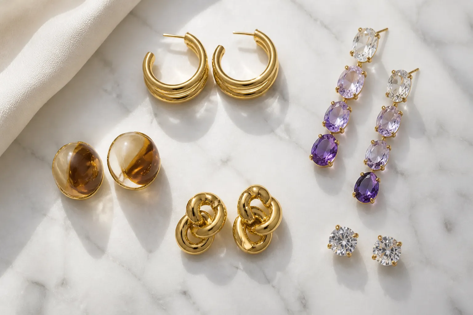 boucle d'oreille or - Flatlay de plusieurs paires de boucles d'oreilles tendance sur fond marbre : créoles plaquées or, pendantes en cristal dégradé, clous en verre bicolore, studs en rhinestone — montrant la diversité esthétique des alternatives
