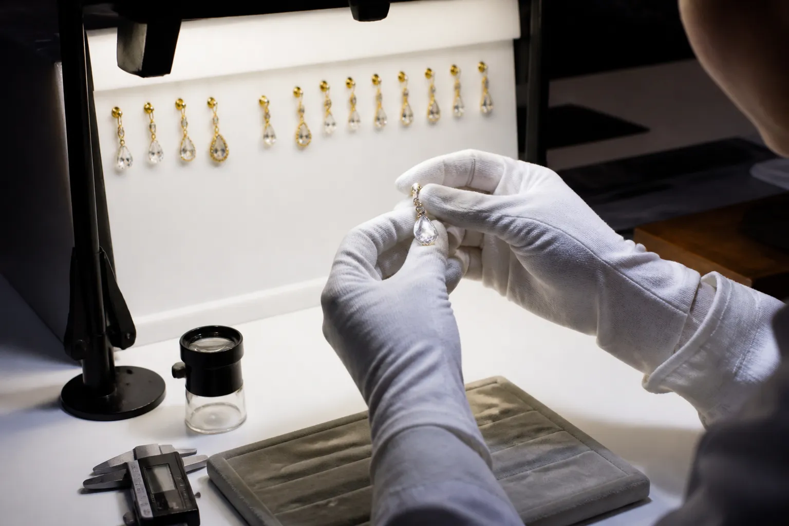 pendientes mujer - Fotografía documental de un espacio de control de calidad en un taller de joyería: mesa de luz iluminada con pendientes mujer colgantes alineados en fila, lupa de mesa, calibre digital y formulario de inspección QC a un lado, manos de técnico con guantes blancos examinando un colgante de cristal, luz de laboratorio cenital uniforme