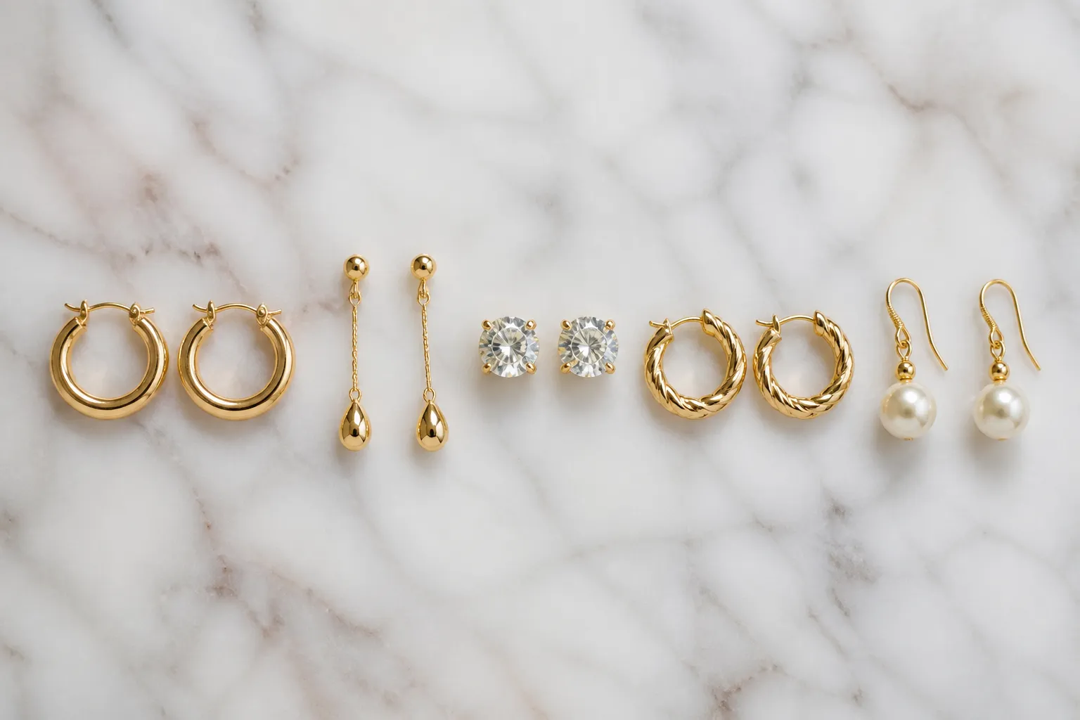 gold ohrringe damen - Flachbild-Arrangement verschiedener Damendohrringe nebeneinander – von Echtgold über vergoldet bis hin zu Kristall- und Glasperlenmodellen, auf einer weißen Marmorfläche fotografiert