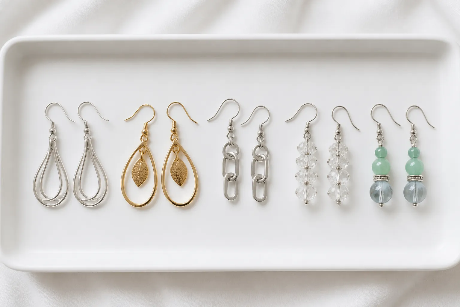 silber ohrringe - Flachansicht verschiedener Ohrring-Materialien – silber, vergoldet, Edelstahl, Kristall, Glasperlen – nebeneinander auf weißem Tabletthintergrund, beschriftet mit Materialname und Preisspanne