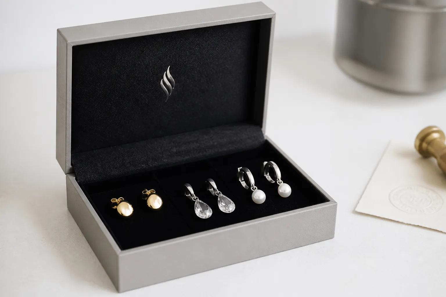 silber ohrringe - Seitenansicht einer geöffneten OEM-Schmuckverpackung mit mehreren Ohrringen-Varianten (vergoldet, Kristall, Perlen) auf schwarzem Samteinleger, Branding-Label sichtbar, Qualitätskontroll-Stempel im Hintergrund