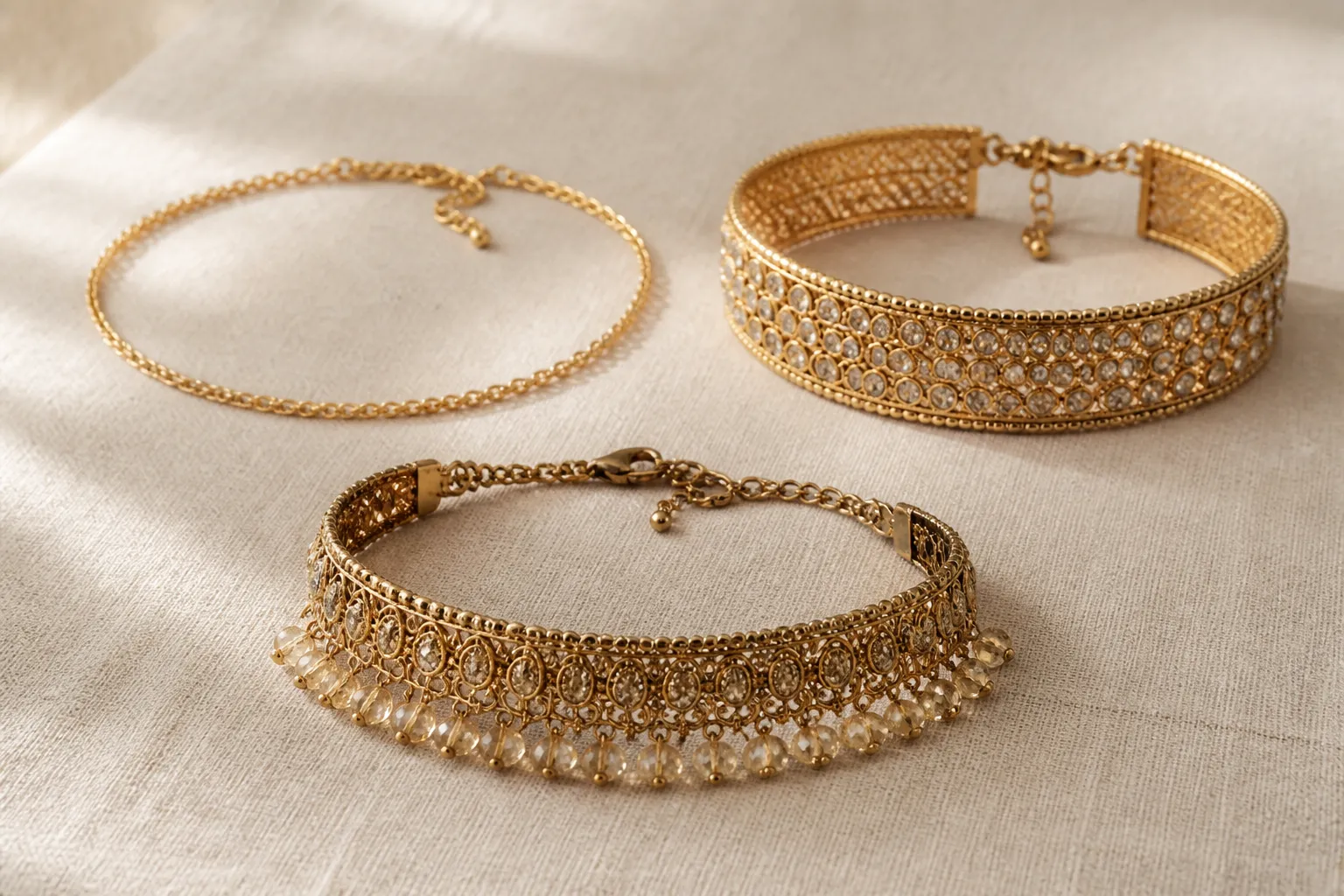خناقه ذهب - Photorealistic close-up of three gold choker necklaces arranged on a neutral cream linen surface — one thin delicate chain, one wide band with crystal embellishments, one vintage-style with glass beads — warm natural studio lighting from the left, each piece showing distinct texture and craftsmanship detail