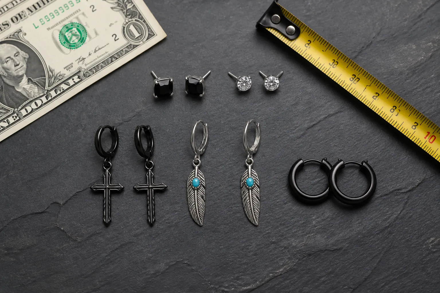 men earrings - Editorial flat-lay of men's stud earrings, drop earrings, and hoop styles arranged on a dark textured surface with a U.S. dollar bill and a tape measure beside them, illustrating scale and local market context