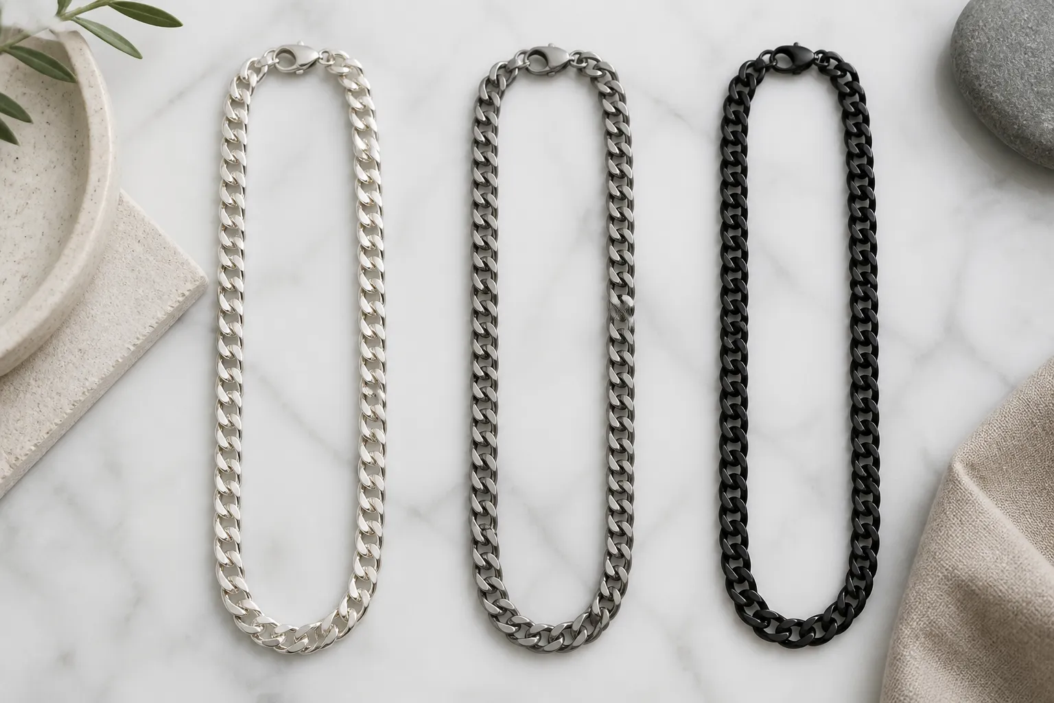 chaine argent homme - Photographie éditoriale comparant côte à côte trois chaînes pour homme — une en argent 925 poli, une en acier inoxydable brossé et une en titane noir — posées sur fond de marbre blanc pour illustrer les différences visuelles de finition