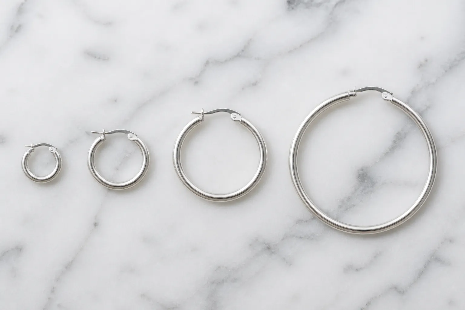 pendientes aro plata - Fotografía editorial con fondo neutro mostrando una selección de pendientes aro plata en diferentes diámetros —10 mm, 25 mm, 40 mm y 60 mm— sobre superficie de mármol blanco, con etiqueta de peso y ley de la plata visible