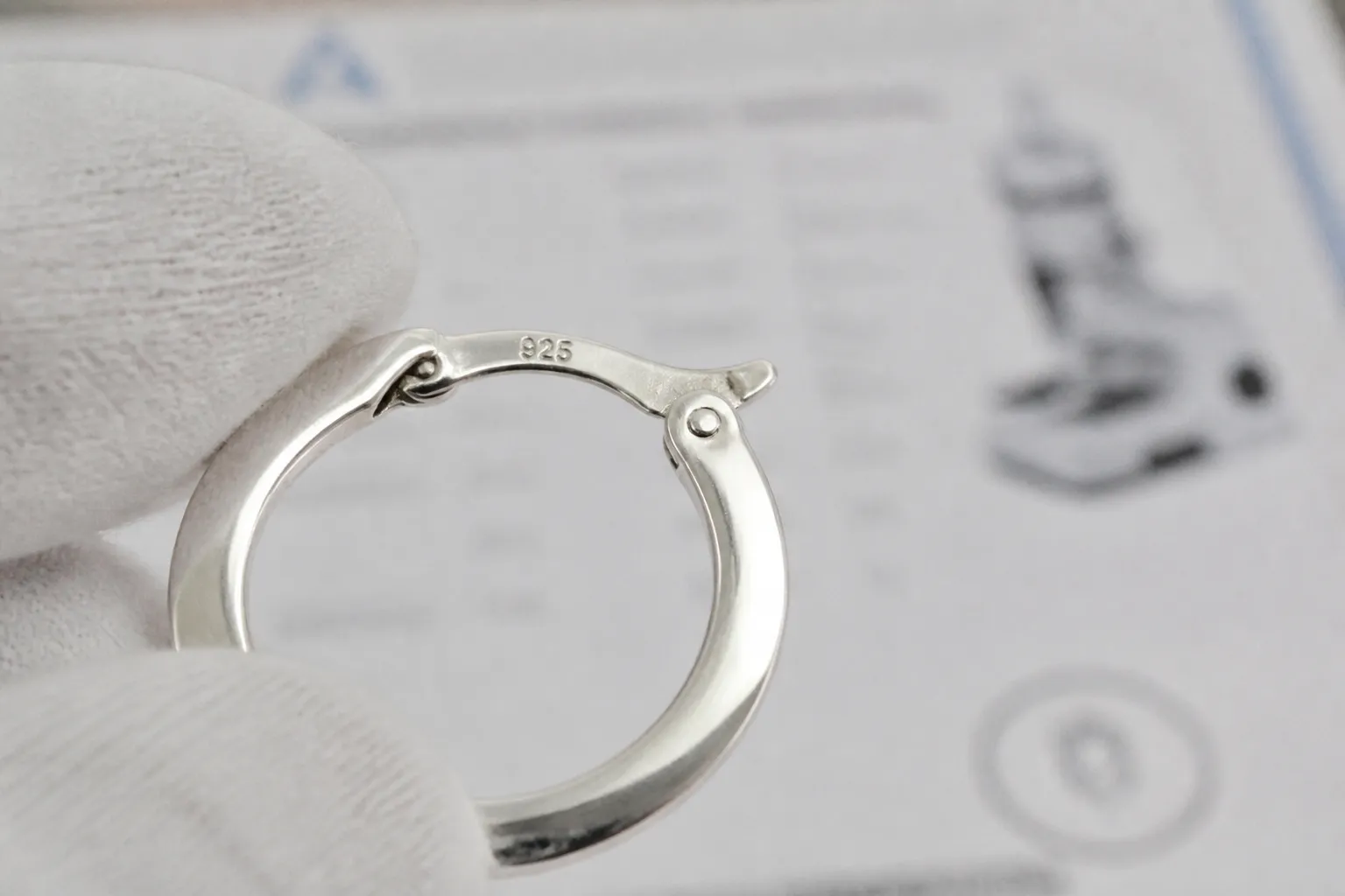 pendientes aro plata - Fotografía de producto profesional mostrando detalle en primer plano del mecanismo de cierre de un pendiente aro plata 925 con sello de ley grabado visible, junto a un certificado de análisis de laboratorio parcialmente desenfocado en el fondo