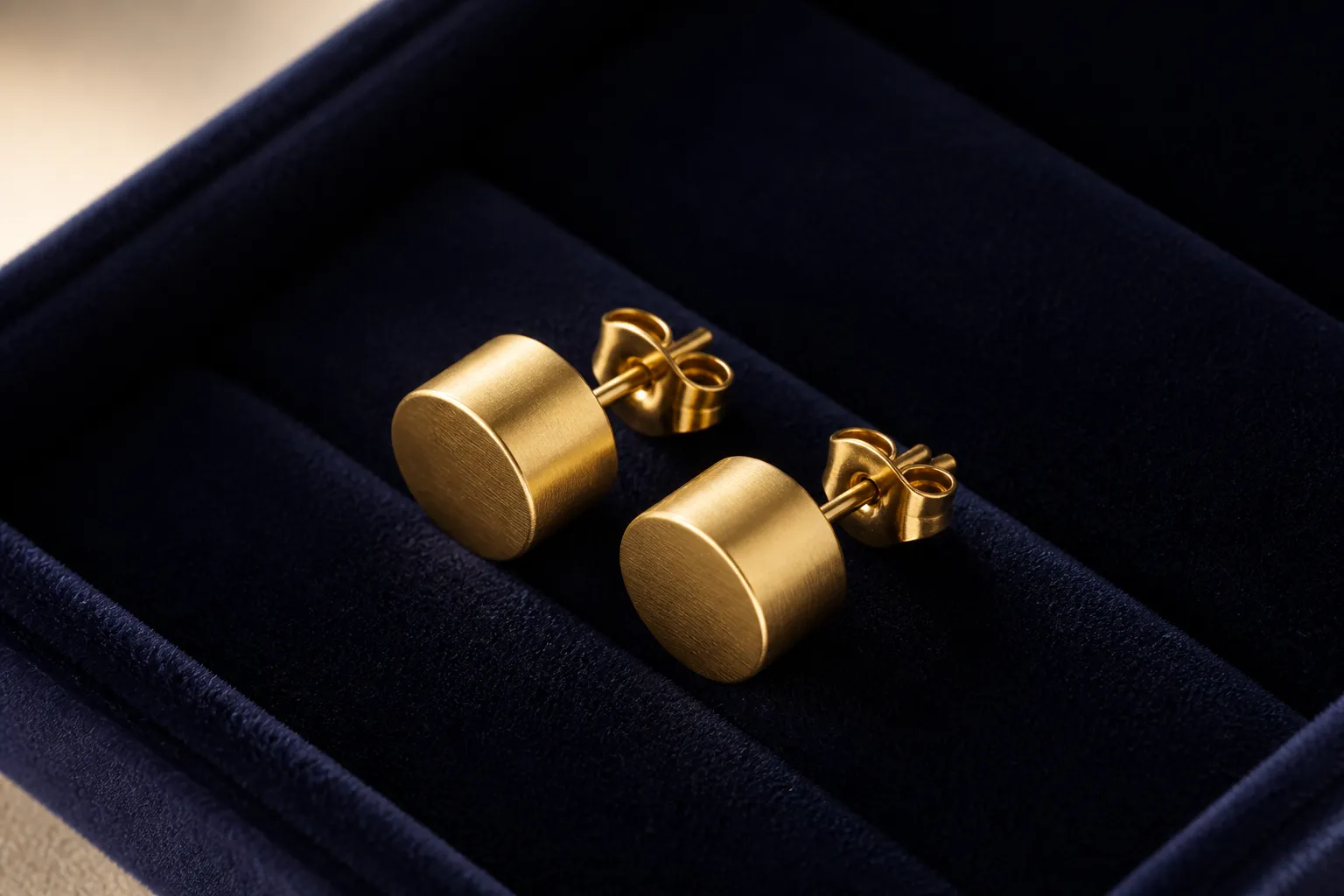 طبلة ذهب - Photorealistic close-up of a pair of gold cylindrical drum earrings (طبلة ذهب) displayed on a dark velvet jewelry tray, warm studio light from the side highlighting the metallic texture and geometric form, documentary product photography style, no text