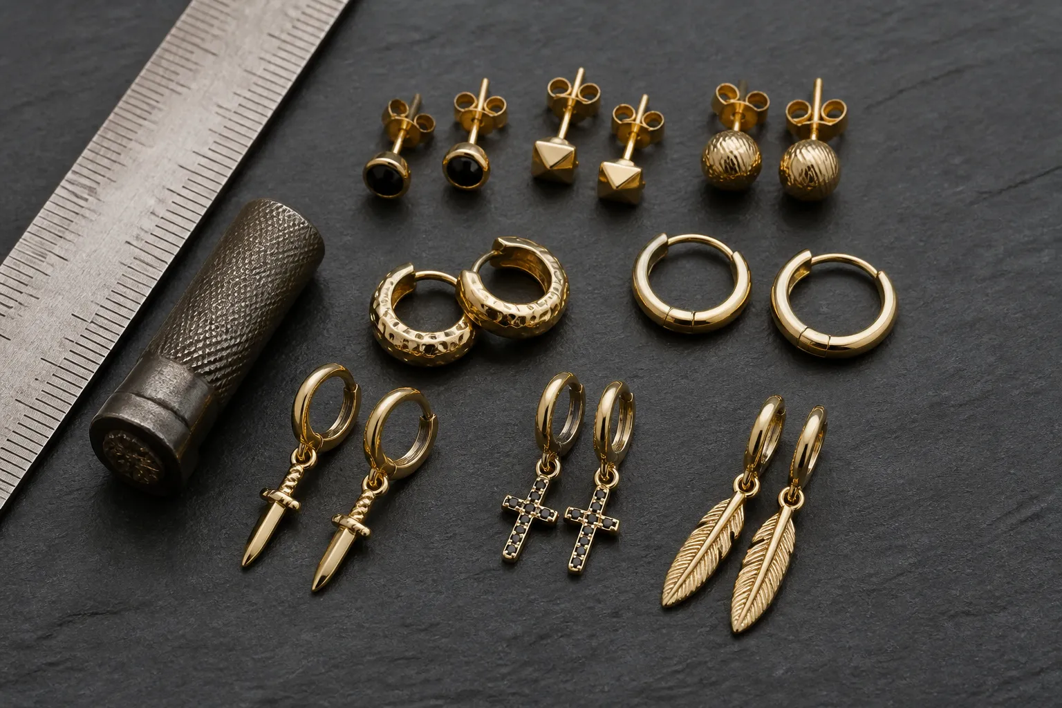 gold earrings for men - A flat-lay editorial photo of assorted gold earrings for men — studs, small hoops, and drop styles — arranged on a dark textured surface with a ruler and hallmark stamp nearby, illustrating the variety of styles and the quality inspection process relevant to cross-border buyers