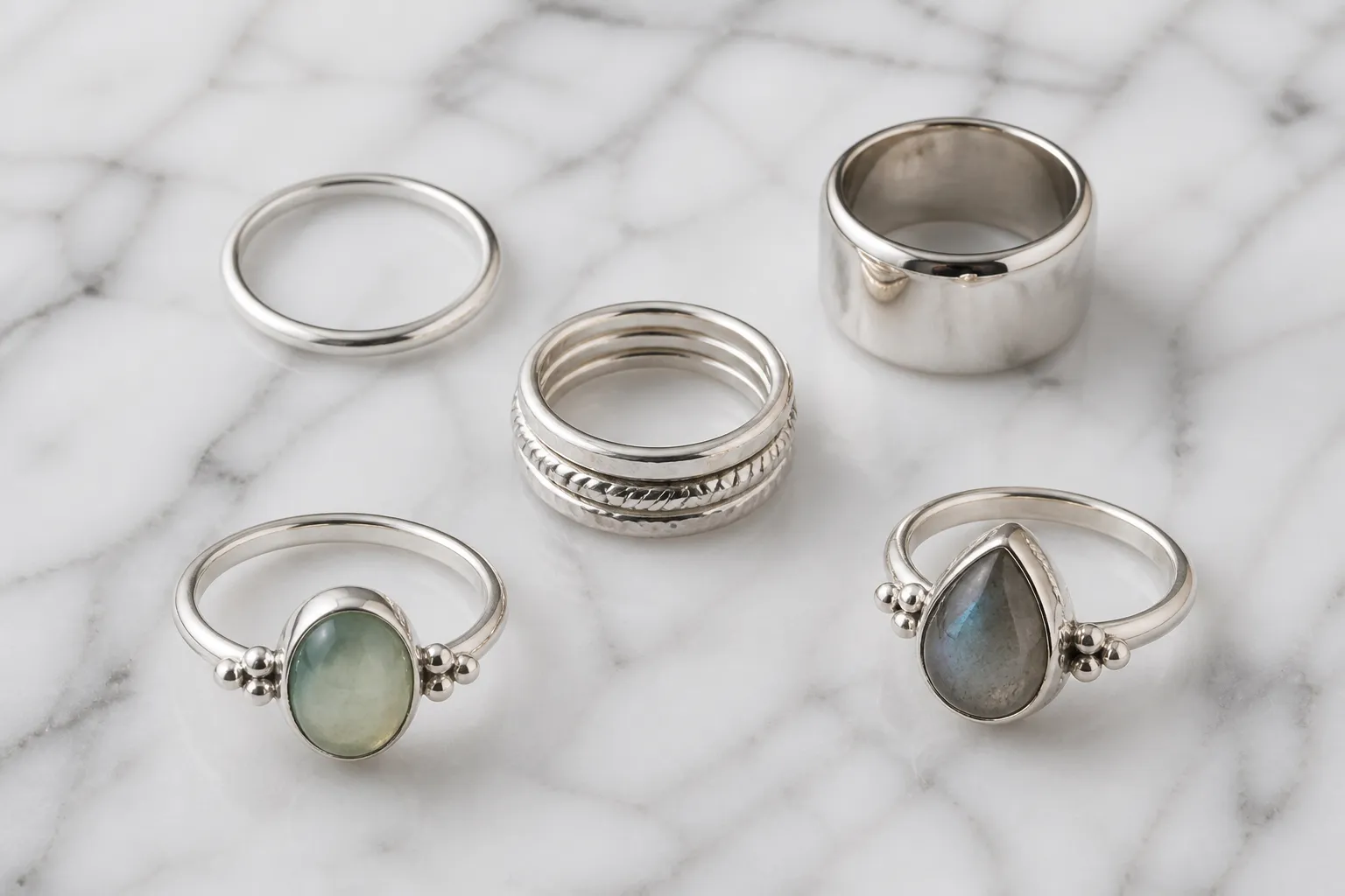 anillos plata - Fotografía editorial de una selección de anillos plata de distintos estilos —minimalistas, con piedras, apilables y de banda ancha— sobre una superficie de mármol blanco, mostrando el rango de opciones disponibles para compradores B2B y DTC