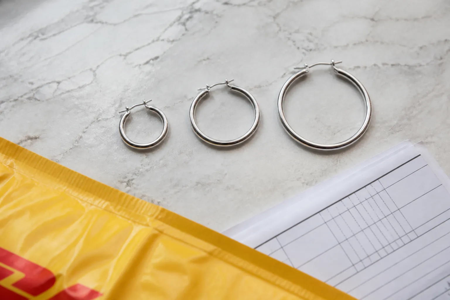 white gold hoop earrings - Photorealistic flatlay of white gold hoop earrings in graduated sizes (small, medium, large) arranged on a textured white marble surface alongside a DHL shipping label and a customs declaration form — illustrating the cross-border sourcing reality