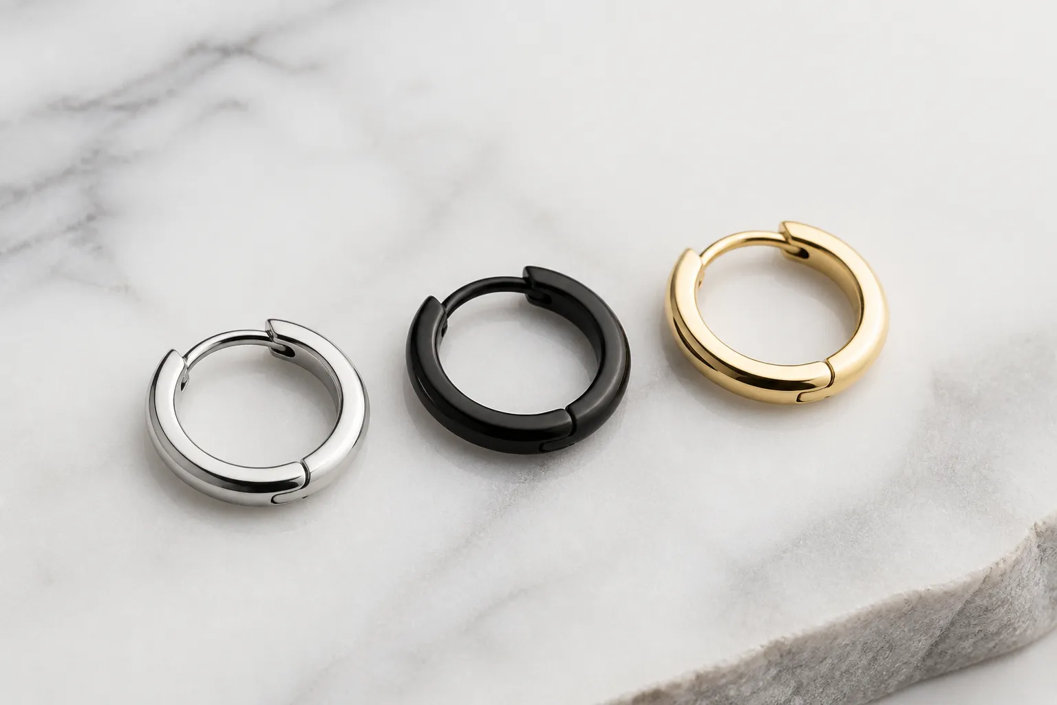 pendientes aro hombre - Selección editorial de pendientes aro hombre en distintos acabados — plata, acero negro y oro — dispuestos sobre una superficie de mármol blanco con iluminación de estudio