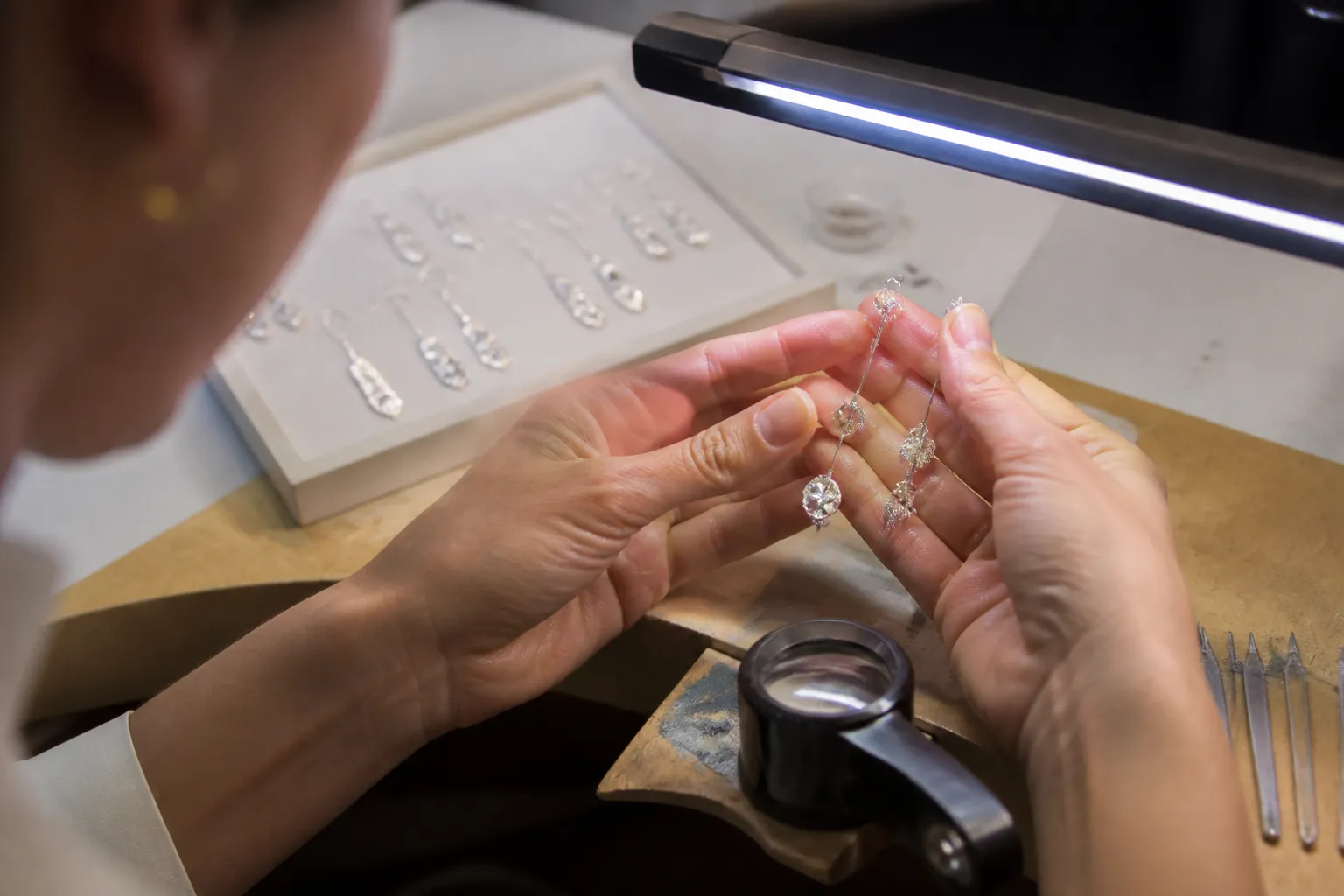 pendientes largos plata - Foto de proceso de control de calidad en taller: inspector examinando pendientes largos colgantes bajo luz LED, con lupa y tabla de muestras aprobadas al fondo
