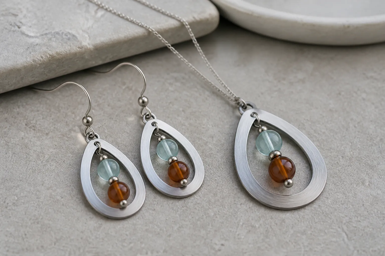 edelstahl schmuck - Nahaufnahme eines Sets aus Edelstahl-Ohrhängern und einer Halskette mit zweifarbigem Glasperlen-Design auf neutralem Hintergrund, die Oberflächenqualität und Finish deutlich zeigend