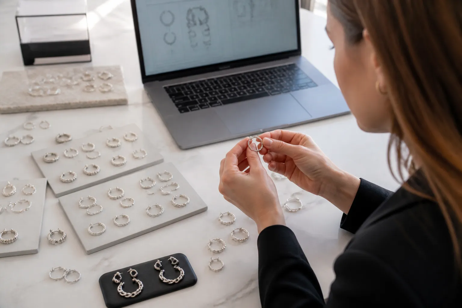 pendientes de plata mujer - Foto editorial de una compradora profesional revisando muestras de pendientes de plata mujer sobre una mesa de trabajo, con un portátil abierto mostrando una hoja de especificaciones técnicas en segundo plano