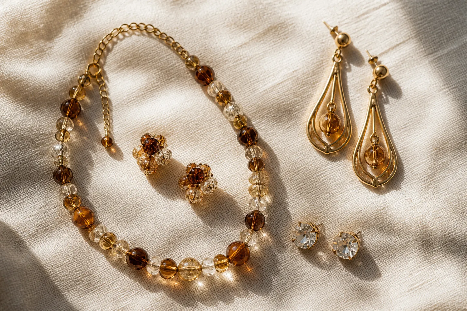 محلات ذهب بني ياس - Annotated product mockup of a STRAND jewelry collection display: a flat-lay top-down shot of drop earrings, rhinestone studs, and a vintage two-tone glass bead necklace-earring set arranged on a cream linen fabric, with small Arabic and English labels indicating material and style — warm natural window light, commercial photography feel