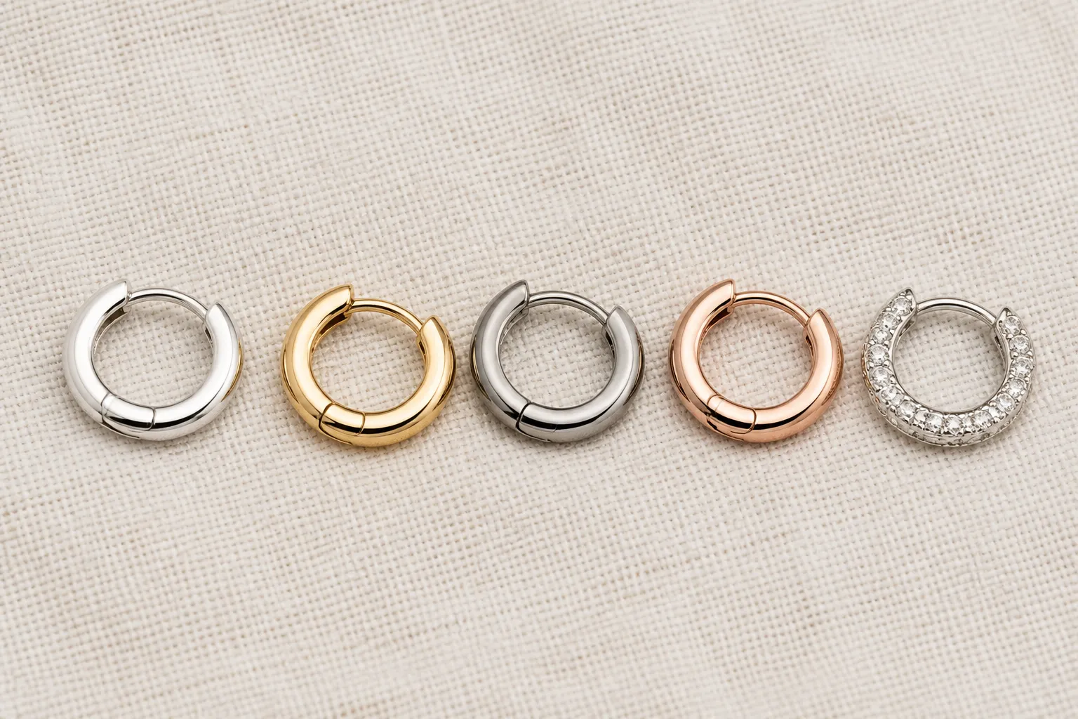 ohrringe creolen silber - Flache Aufsicht auf verschiedene Creolen-Stile nebeneinander – Silber, vergoldet, Edelstahl, Roségold und mit Kristallbesatz – auf neutralem Leinenhintergrund, typische Produktfotografie für B2B-Kataloganfragen
