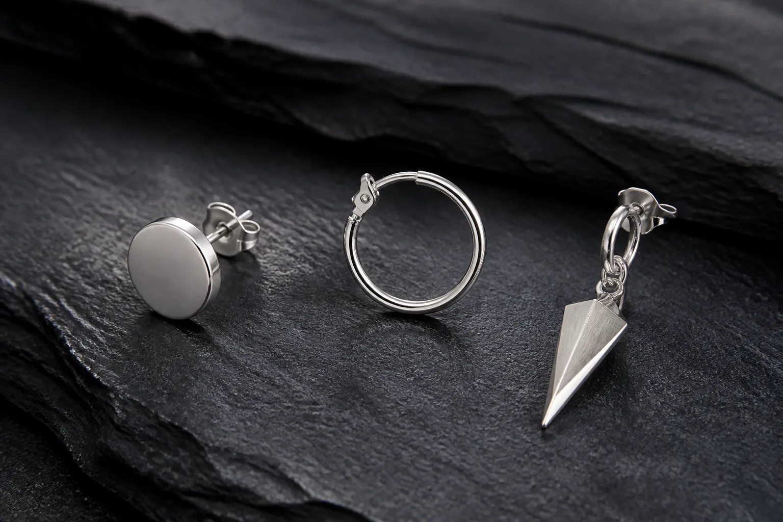 pendientes plata hombre - Fotografía editorial realista de tres estilos distintos de pendientes de plata para hombre —un stud de círculo liso, un aro fino y un colgante geométrico— dispuestos sobre una superficie de pizarra oscura, iluminación lateral suave que resalta los acabados metálicos, sin texto en la imagen