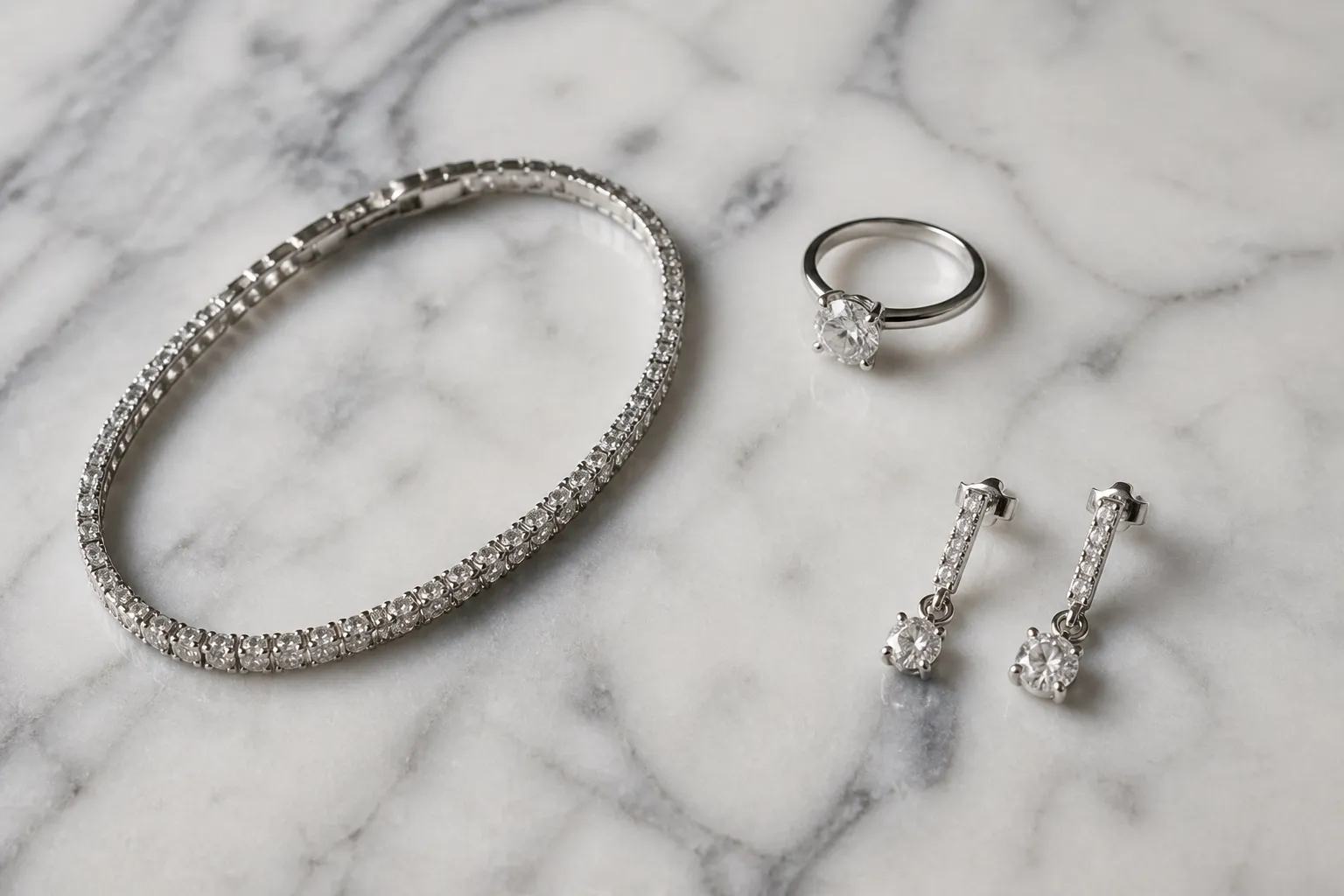 ذهب أبيض - Photorealistic overhead shot of white gold jewelry pieces — a bracelet, a ring, and drop earrings — arranged on a pale marble surface under soft natural side-lighting, showing the rhodium-plated silver sheen against the stone texture, no text overlays