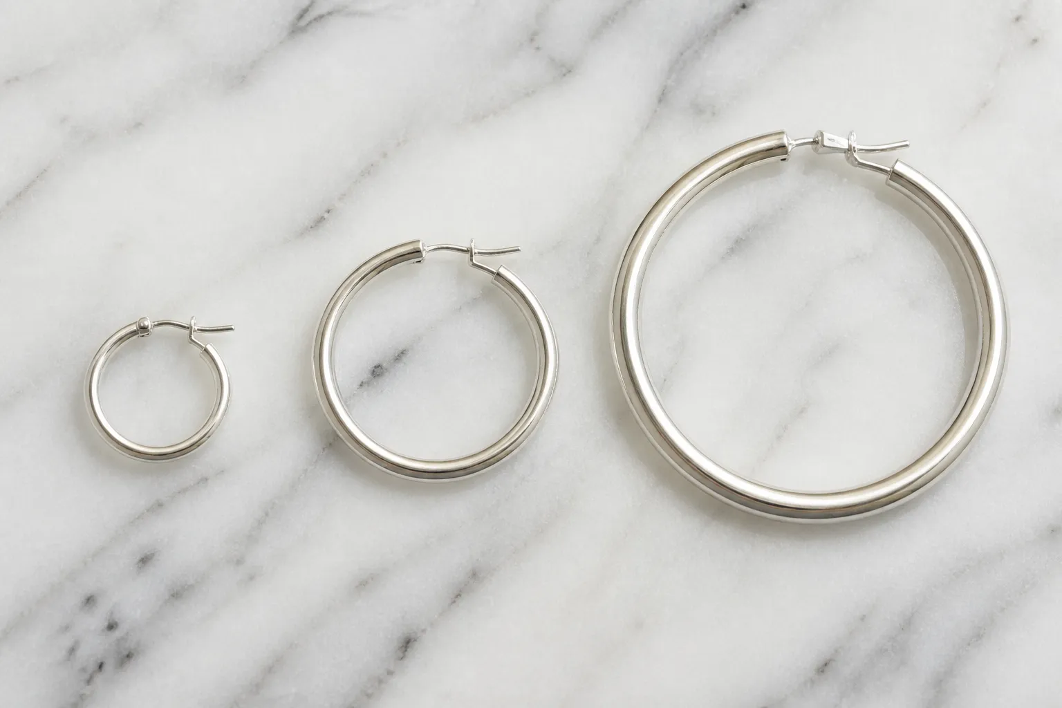 pendientes aro de plata - Fotografía editorial de varios pendientes aro de plata de distintos diámetros (15mm, 30mm, 50mm) sobre superficie de mármol blanco, mostrando diferencias de grosor de aro y tipo de cierre