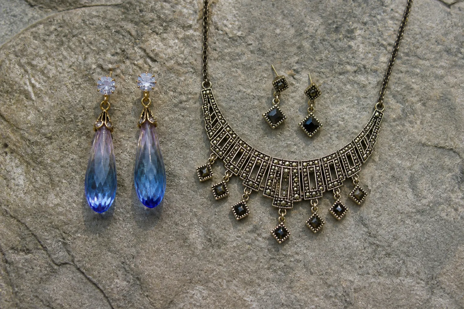 custom jewelry - Editorial flatlay of a gradient crystal drop earring set alongside a vintage geometric necklace and earring set, styled on a stone surface — showing the aesthetic range achievable through stock catalog sourcing without custom production investment