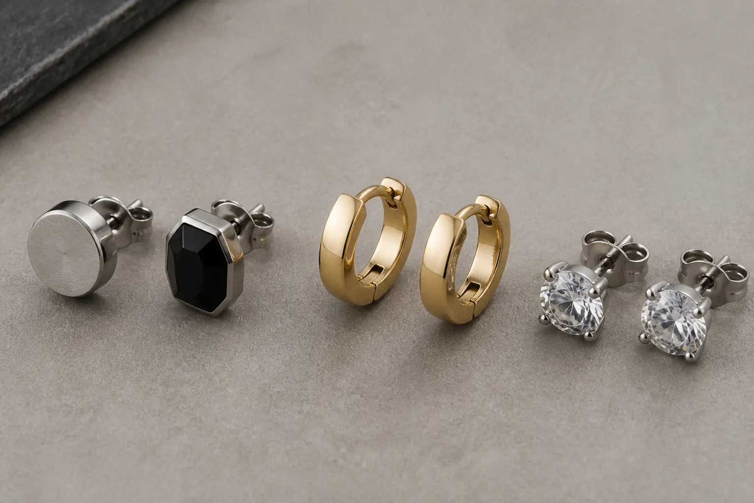 herren ohrringe - Flachaufnahme verschiedener Herren Ohrringe nebeneinander – Edelstahl-Studs, vergoldete Hoops und Silber-Zirkonia-Stecker auf neutralem Hintergrund, Materialbeschriftungen als Overlay