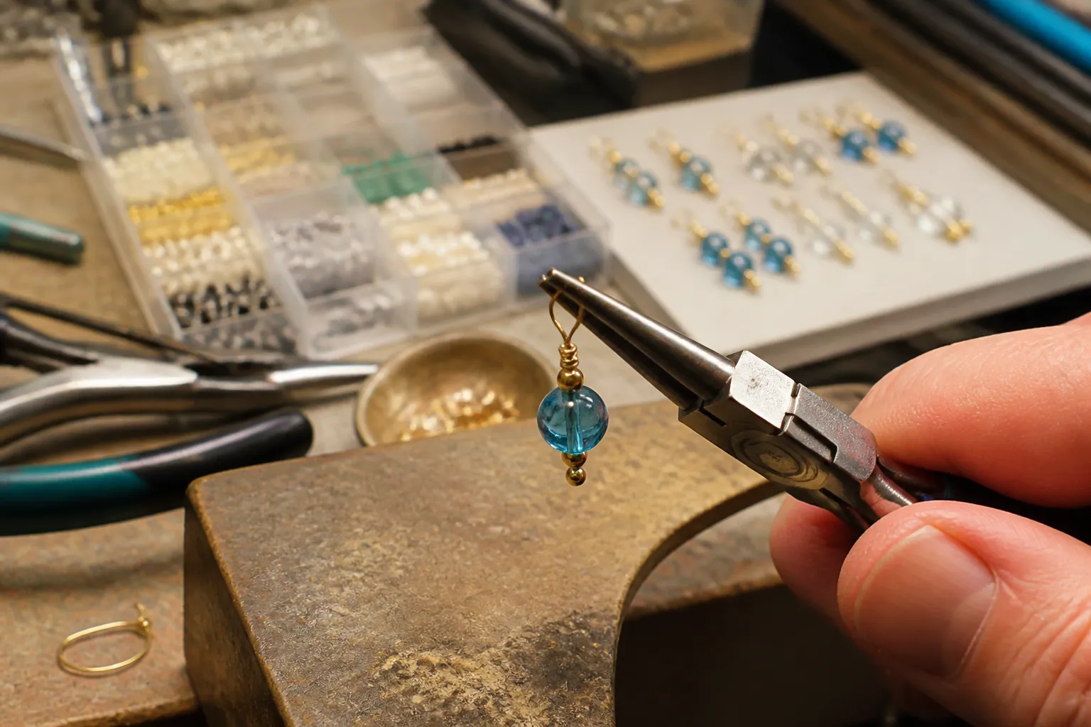 boucles d'oreilles pendantes - Zoom sur le détail d'assemblage d'une boucle d'oreilles pendante en atelier — tige à œil sur perle de verre, pince à bijoux visible, poste de contrôle qualité en fond