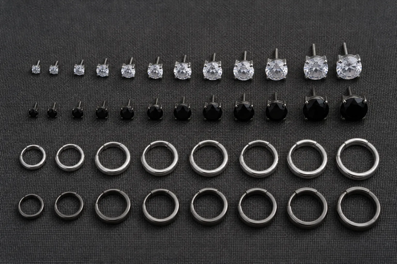 ohrringe herren - Flache Produktaufnahme verschiedener Herrenohrringe — Ohrstecker in 3 mm bis 7 mm mit weißen und schwarzen Zirkonia-Steinen, Kreolen aus Silber und Stahl, angeordnet auf strukturiertem dunkelgrauem Hintergrund, mit Materialbezeichnungen beschriftet