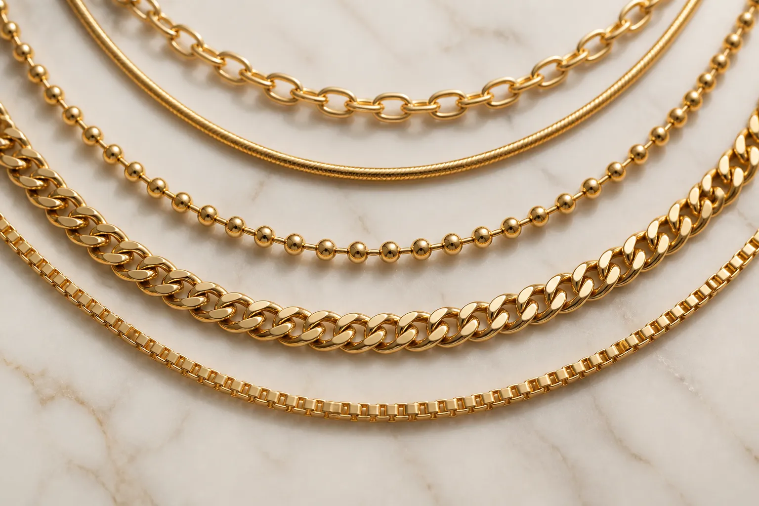 chaine en or - Photorealistic flat-lay of five different gold chain necklaces arranged in an arc on a matte white marble surface — forçat, serpentine, maille bille, gourmette and veneziana styles visible, warm studio lighting from above, no text or labels
