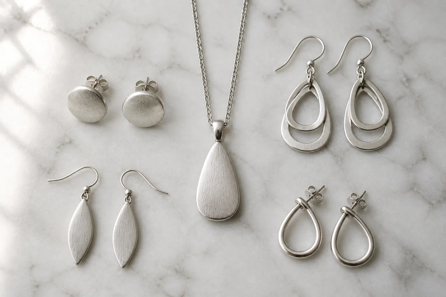 schmuck silber - Photorealistisches Flat-Lay-Foto von sechs verschiedenen Silberschmuckstücken – Ohrstecker, Tropfenohrringe und ein Halskettenanhänger – auf einer strukturierten Marmorunterlage, natürliches Fensterlicht von links, neutrale Draufsicht, klare Detailschärfe auf den Oberflächen