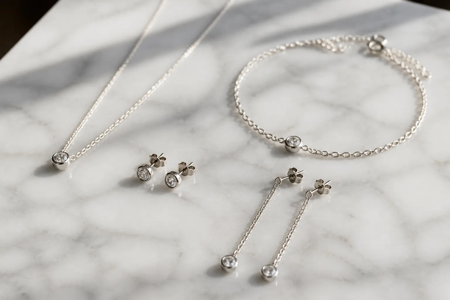 bijoux argent - Photorealistic flat-lay of assorted silver-tone jewelry pieces — minimalist stud earrings, delicate drop earrings, a thin chain necklace and a dainty bracelet — arranged on white Carrara marble, natural side lighting from a window, close-up documentary photography showing surface texture and rhodium plating detail with soft reflections