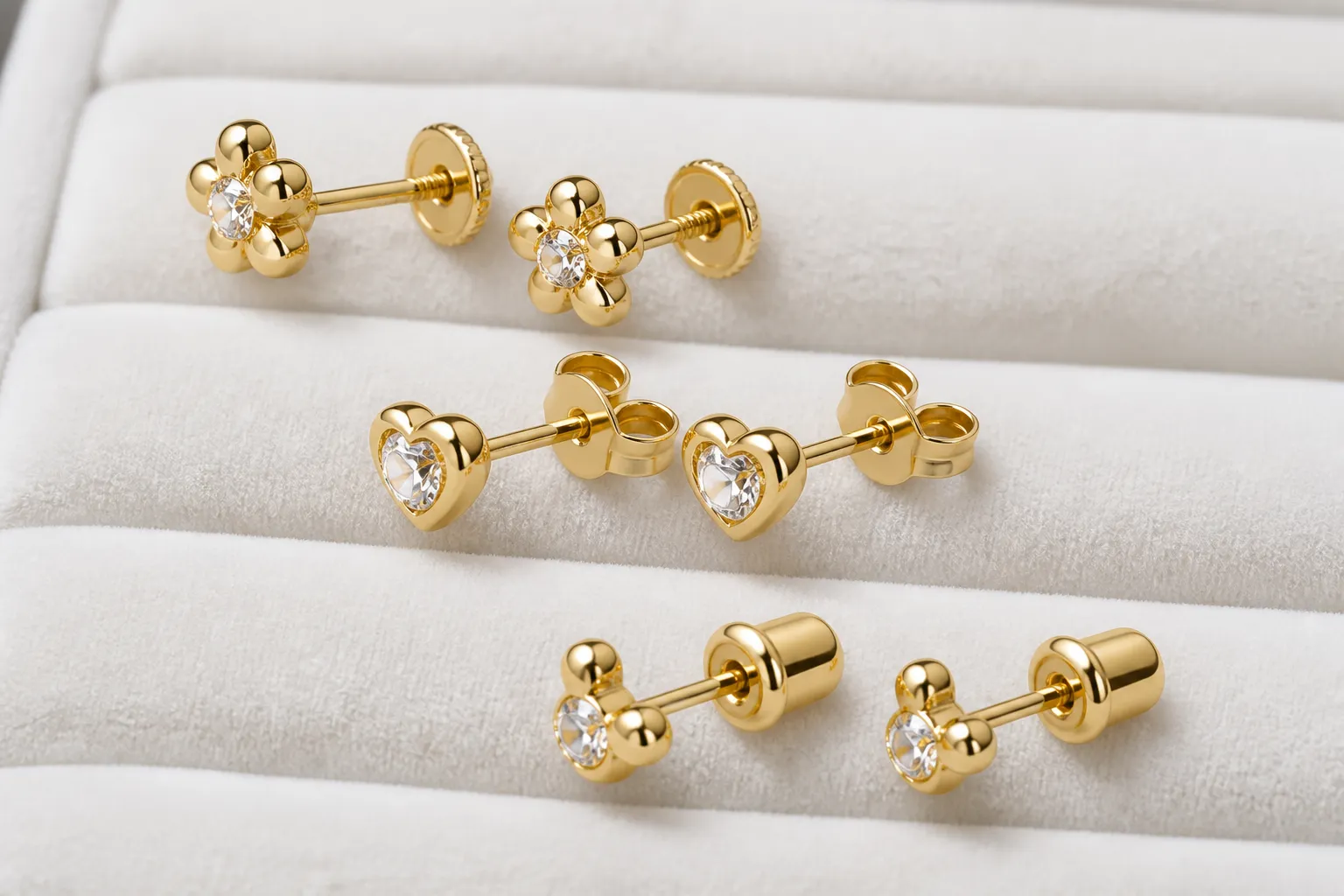 pendientes de bebe oro - Fotografía editorial de cerca mostrando tres pares de pendientes de bebé en oro 18k sobre terciopelo blanco, con distintos tipos de cierre identificados con etiquetas: cierre de rosca, mariposa de seguridad y palomita con tope