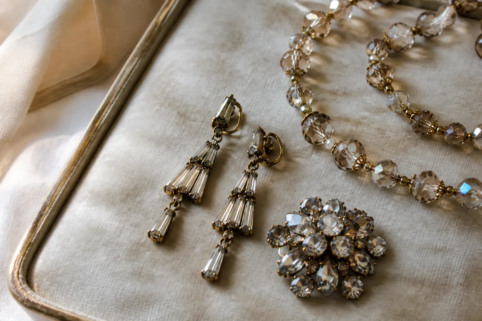 bijoux anciens - Photorealistic flat-lay photography of antique-style jewelry pieces — gold-plated drop earrings with baguette crystals, a glass bead necklace and a rhinestone brooch — arranged on aged ivory velvet fabric, natural window light from the left, shallow depth of field highlighting metal texture and stone facets, no text or overlay