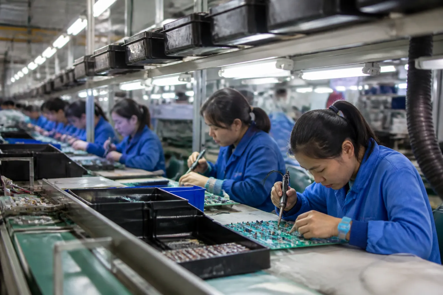 production meaning - 中国电子装配线的逼真广角镜头——身穿蓝色制服的工人在发光的工作站焊接PCB板,头顶传送系统运输零件托盘,明亮的荧光灯工厂照明,真实的工业纪录片风格