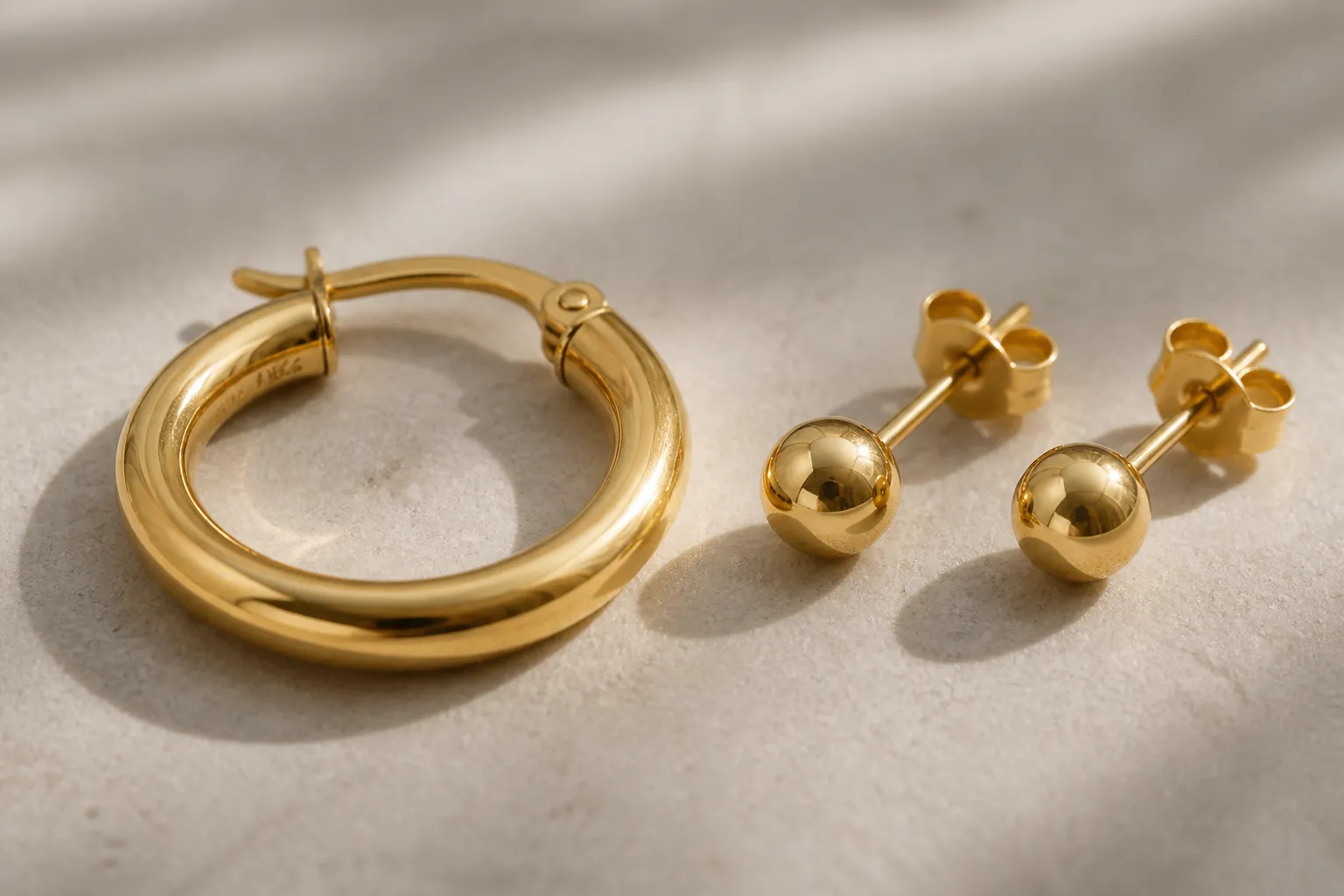 pendientes oro 18k - Primer plano de pendientes tipo aro y botón fabricados en oro amarillo 18k, con el marcado de contraste visible en el cierre, sobre fondo neutro de piedra