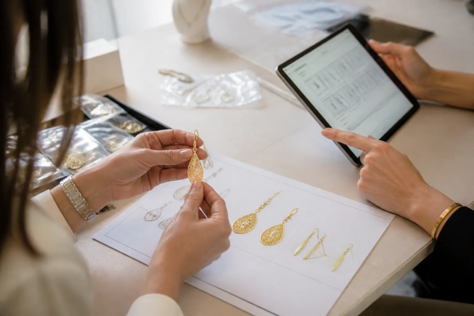 custom jewelry - Editorial photo showing two procurement professionals reviewing jewelry samples at a light table — one holding a drop earring sample against a spec sheet, the other referencing a tablet showing an order management interface