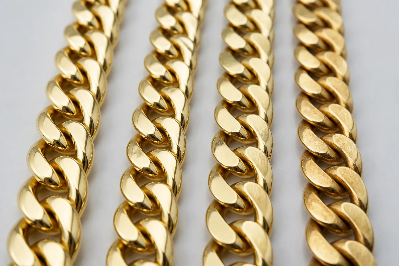 cadena cubana oro - Close-up photograph of several gold Cuban link chains laid side by side on a neutral surface, showing differences in link width, finish quality and material — from solid 18k yellow gold to gold-plated copper and stainless steel