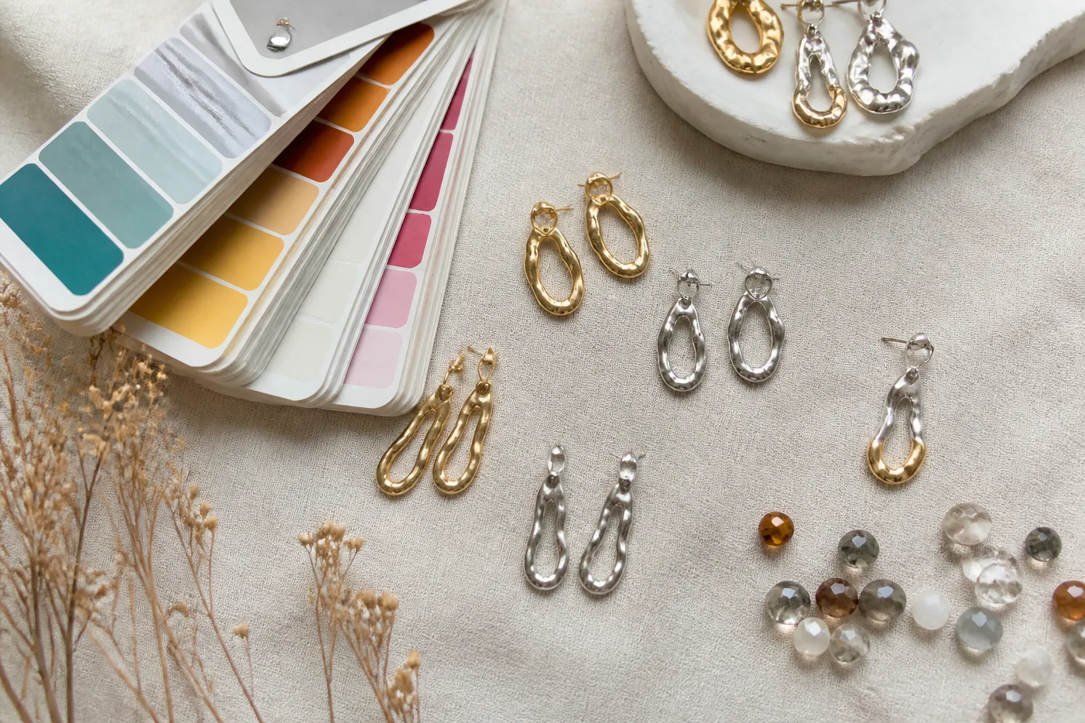 custom jewelry - Photorealistic flatlay showing a jewelry design brief with Pantone color swatches, gold-plated and rhodium-plated copper sample earrings, and glass bead stone selections spread across a procurement review table