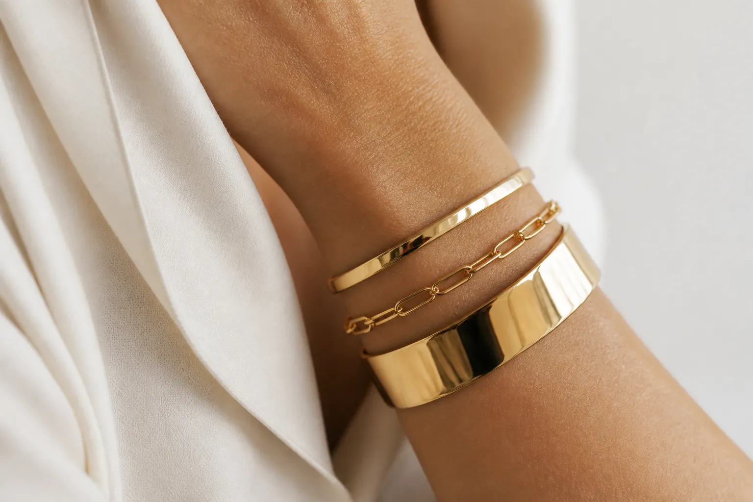 pulsera oro mujer - Fotografía editorial de una mujer con pulseras de oro apiladas en la muñeca — esclava, cadena fina y bangle — sobre fondo neutro blanco, mostrando cómo se combinan diferentes estilos y tipologías en un look coherente