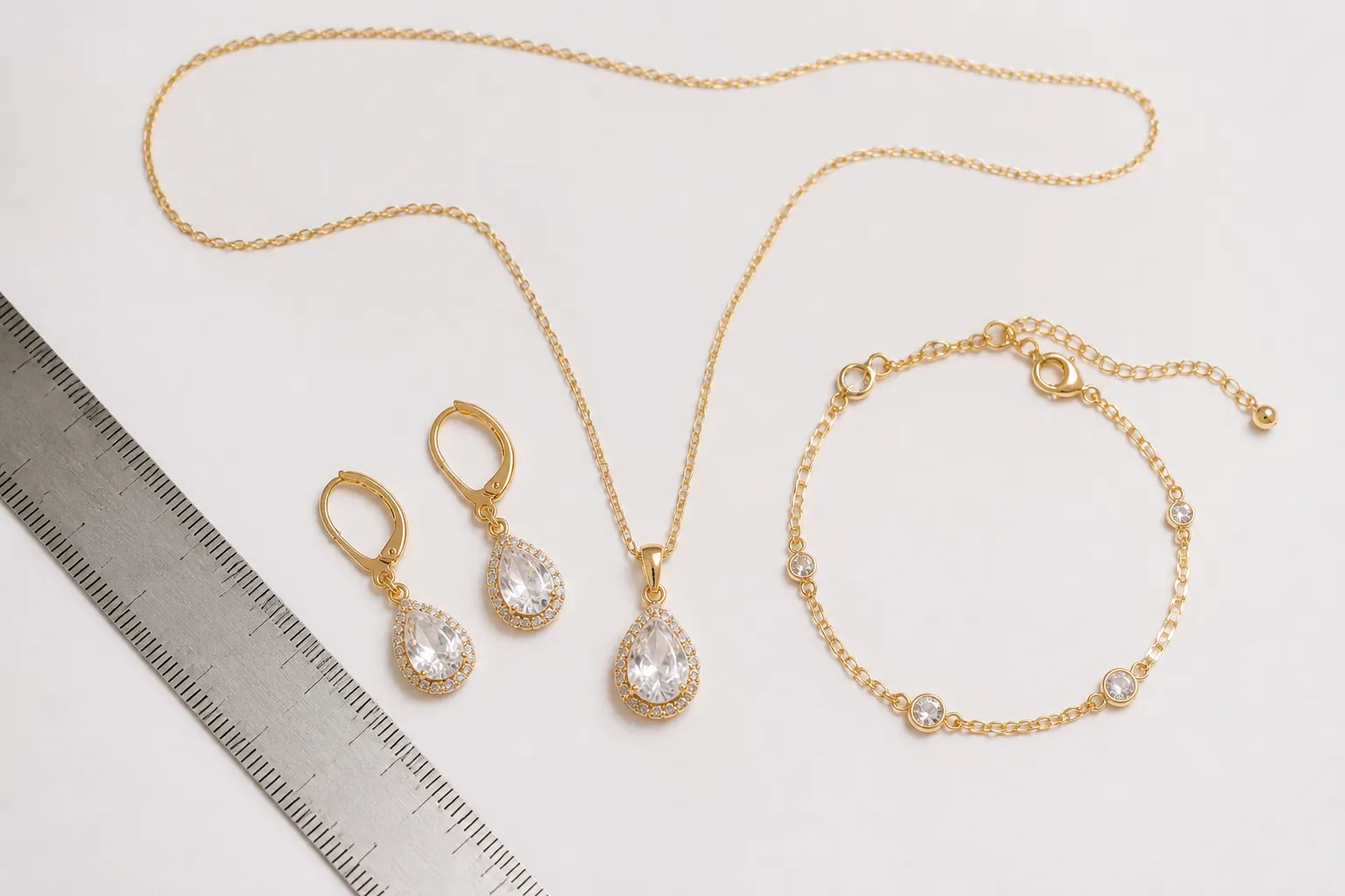 cadena oro mujer - Fotografía de producto plana (flat lay) mostrando una cadena de oro para mujer junto a unos pendientes de gota de cristal y una pulsera a juego, todos en el mismo tono dorado, sobre fondo blanco neutro con escala de longitud en centímetros