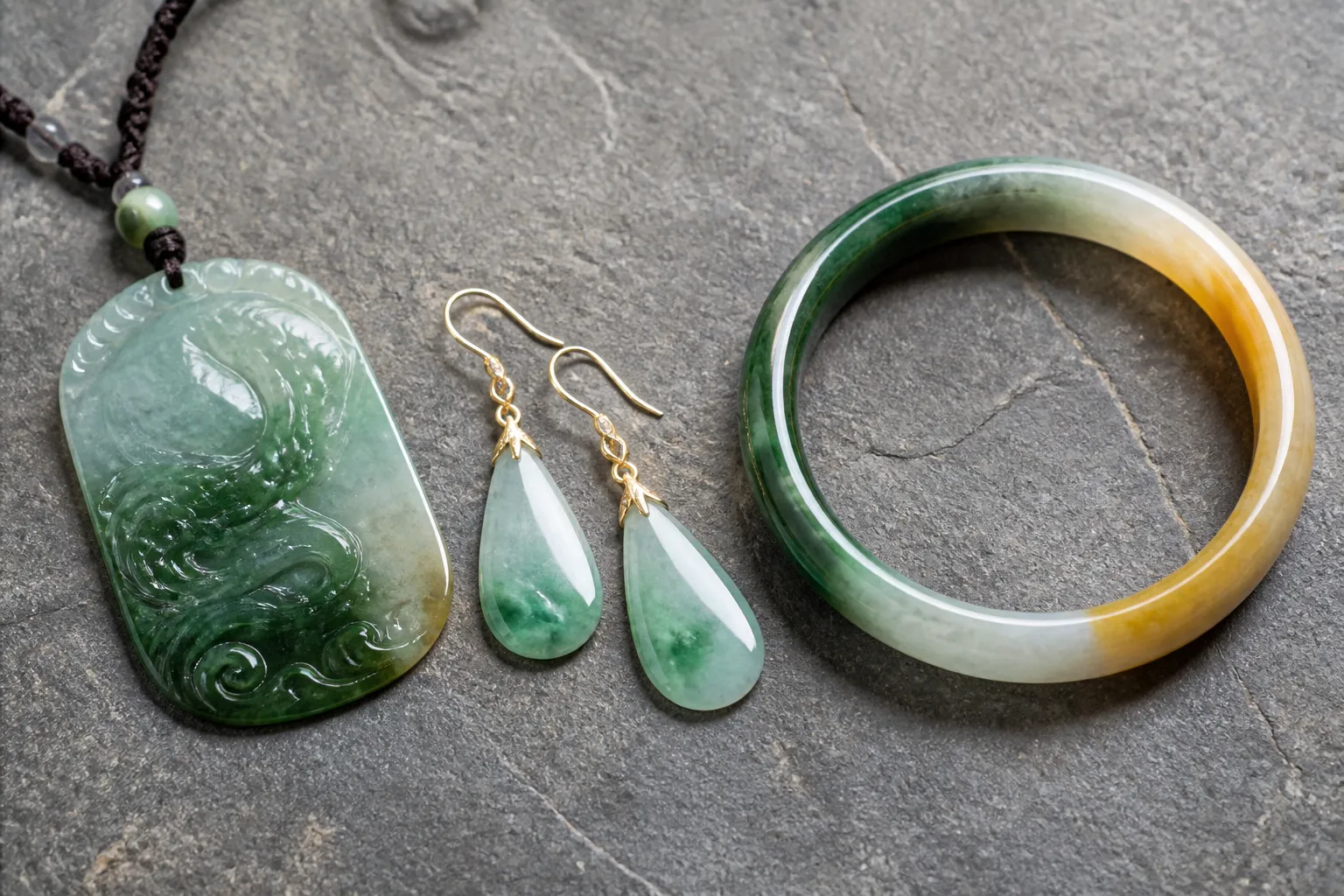jade piedra - Fotografía editorial de piezas de jade piedra natural —colgante, pendientes y pulsera— sobre fondo de piedra gris, mostrando la variación de tonos verdes, blancos y amarillos de la gema en distintos grados de calidad