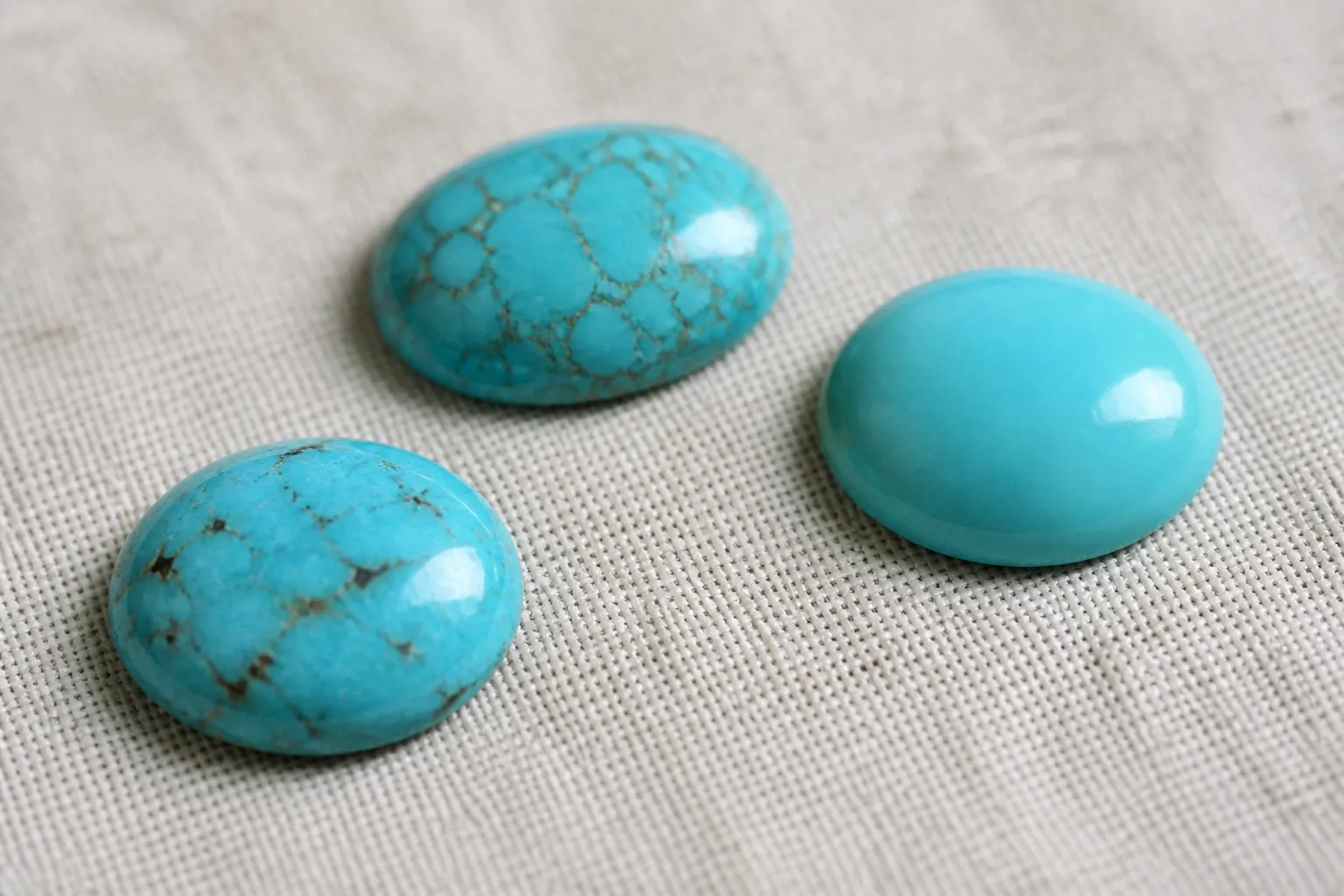 piedra turquesa - Photorealistic macro shot of three distinct turquoise cabochons arranged on a textured linen surface, showing natural matrix veining in the center stone versus stabilized uniform color in the left stone, soft natural window lighting highlighting surface texture differences for buyer comparison
