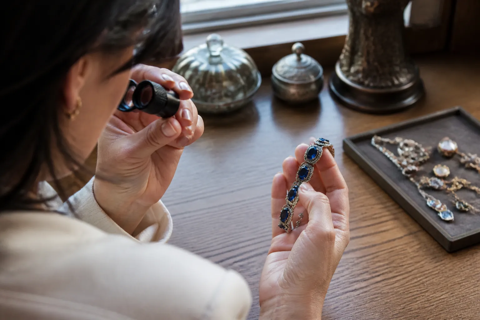 wholesale bracelets - Photorealistic over-the-shoulder shot of a jewelry buyer examining a vintage gemstone bracelet with a loupe on a wooden desk, natural window light illuminating metal texture and stone clarity, shallow depth of field, documentary style