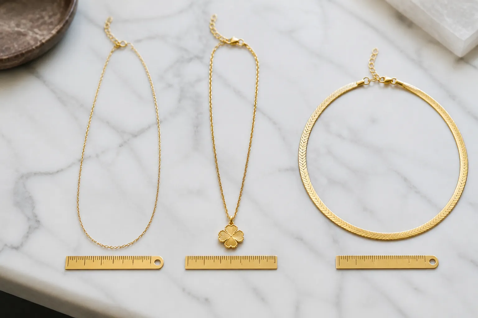 collier en or - Photographie éditoriale d'une sélection de colliers en or — chaîne fine dorée, pendentif trèfle plaqué or et collier ras-du-cou — disposés sur fond de marbre blanc avec étiquettes de longueur