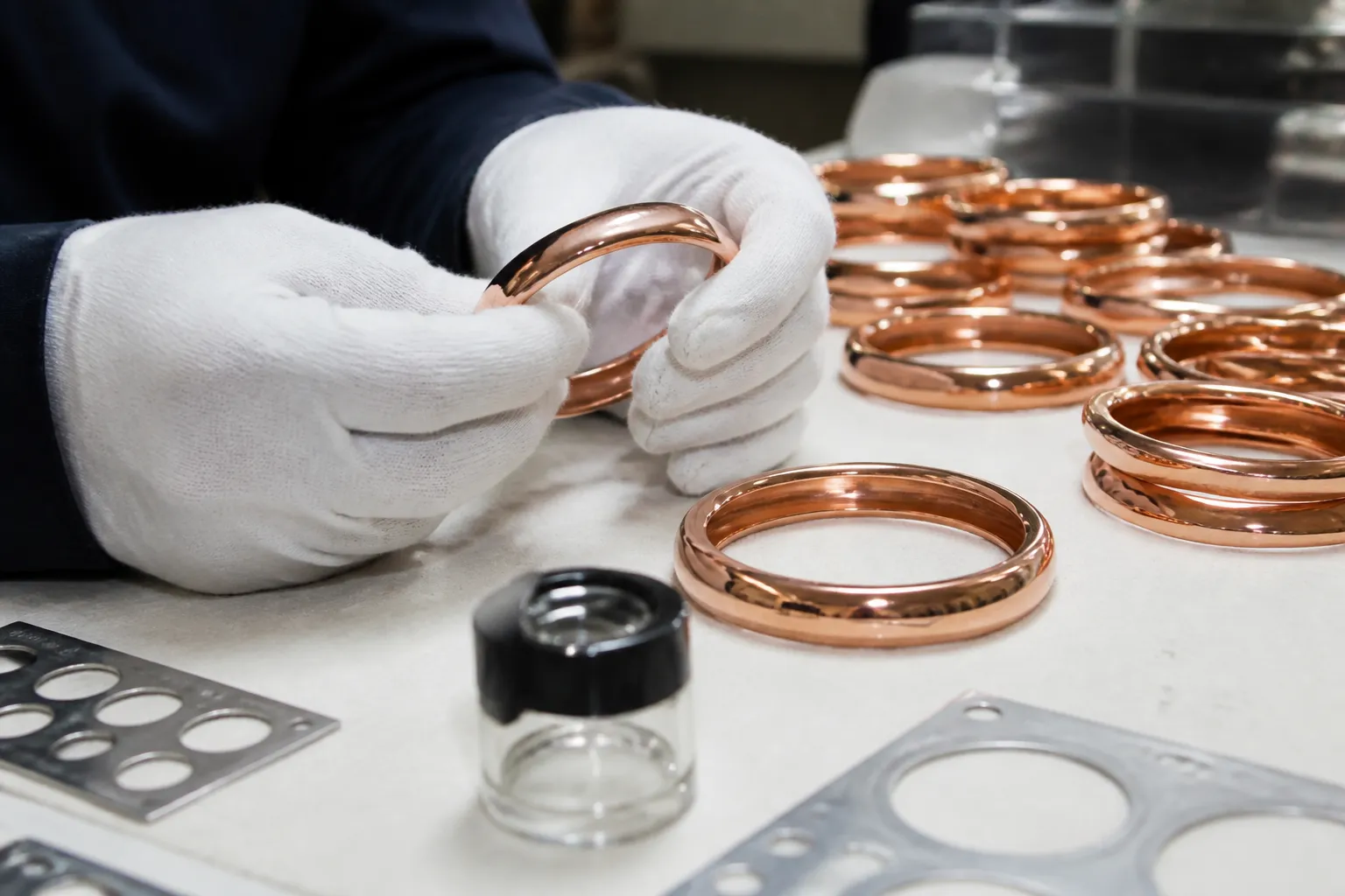 pulsera de cobre - Fotografía de proceso de control de calidad en taller de joyería — manos con guantes inspeccionando pulseras de cobre sobre mesa iluminada, con lupas y plantillas de medición visibles