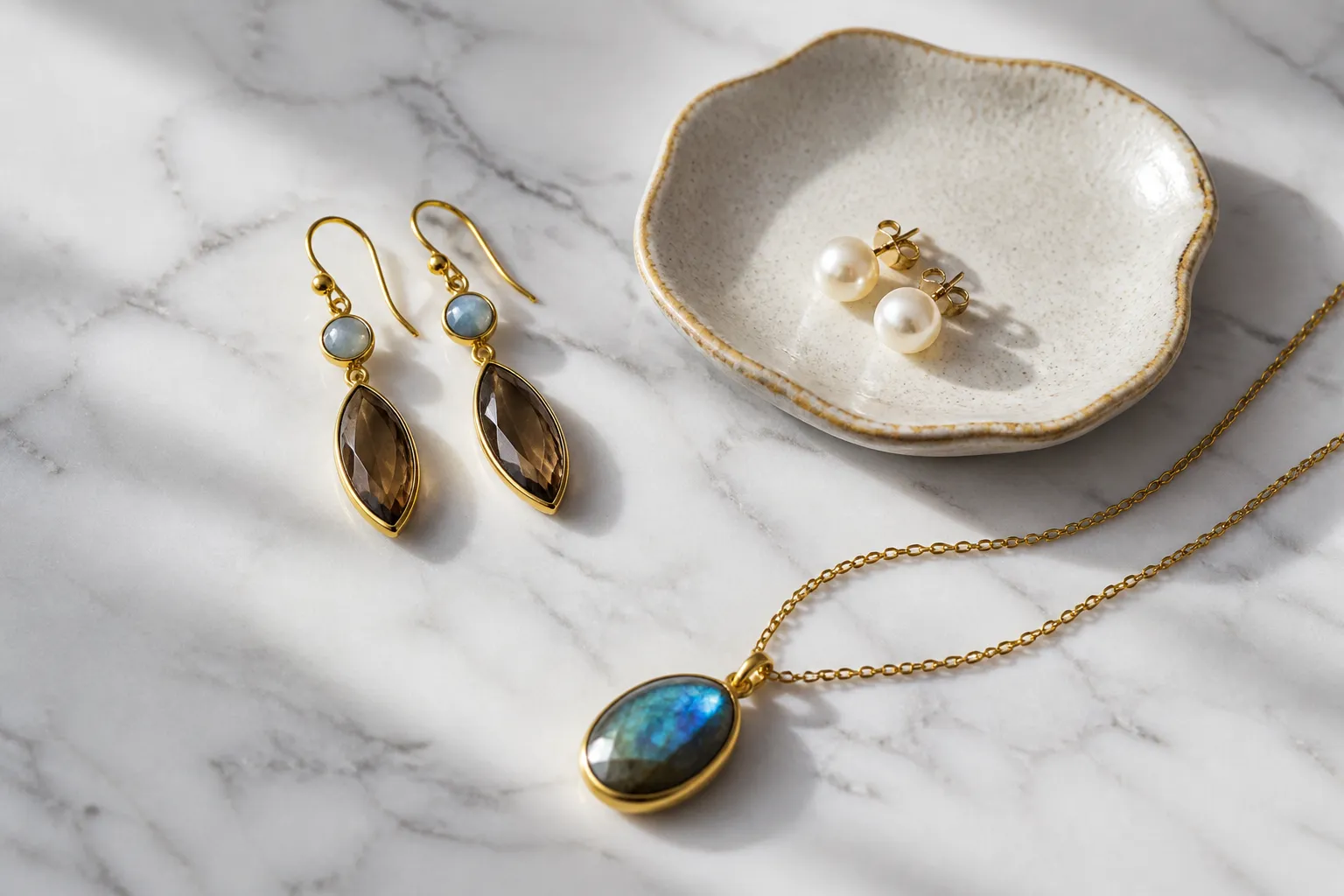 pierre précieuse - Editorial flat-lay on a white marble surface showing three jewelry pieces — a pair of gold-plated drop earrings with natural stone accents, a pearl stud earring pair on a small ceramic tray, and a delicate necklace with a semi-precious stone pendant — arranged in a loose triangle composition with natural side lighting casting soft directional shadows, authentic product photography without text