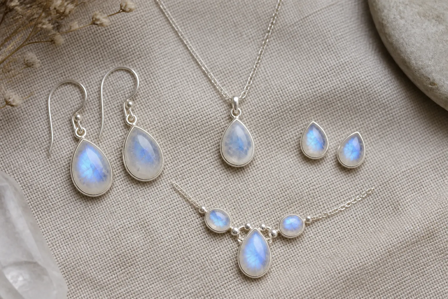 pierre de lune - Flat lay éditorial de plusieurs bijoux en pierre de lune — boucles d'oreilles pendantes, clou, set collier — disposés sur fond de lin naturel, illustrant la polyvalence stylistique de la gemme pour une ligne de bijouterie fantaisie moderne