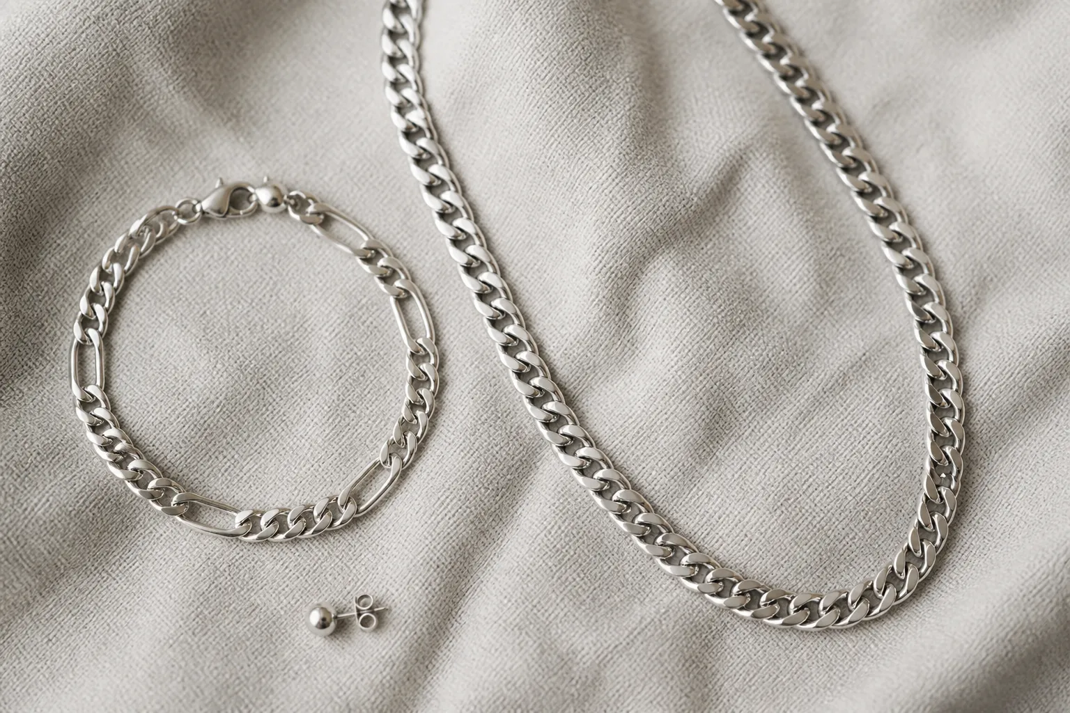 armband männer silber - Flat-lay-Aufnahme eines kuratierten Herren-Schmucksets: silbernes Gliederarmband, Königskette und schlichter Silber-Stecker auf hellem strukturierten Hintergrund — mit Beschriftungen der Materialdetails
